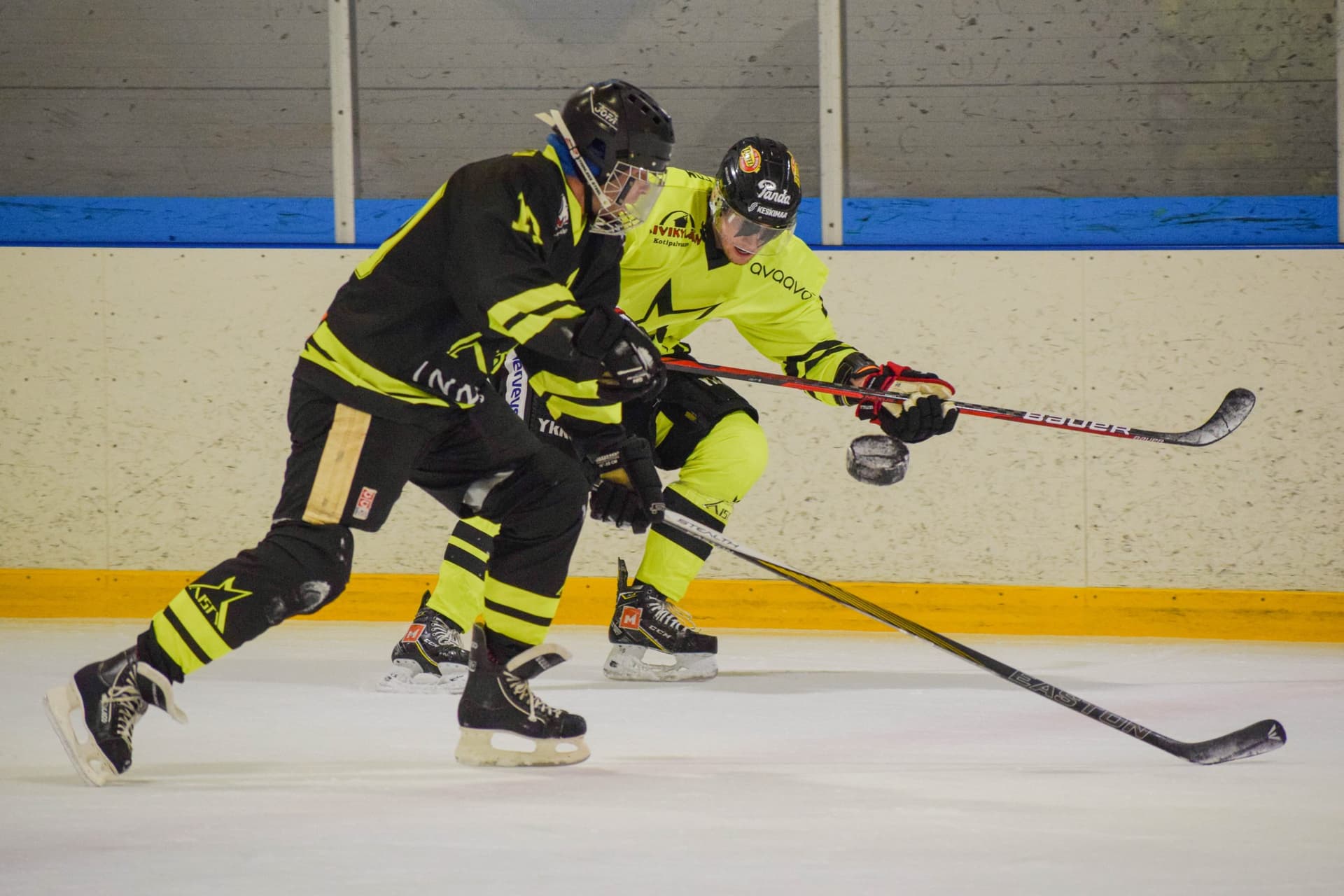 Pelitilannekuva näkövammaisjääkiekko-ottelusta jossa flumino- ja mustapaitaiset Aisti Sportin pelaajat luistelevat vauhdilla kaukalon vierustalla kiekon perässä. Molemmilla pelaajilla on mailat etukenossa ja musta kiekko on ilmassa pelaajien edessä.