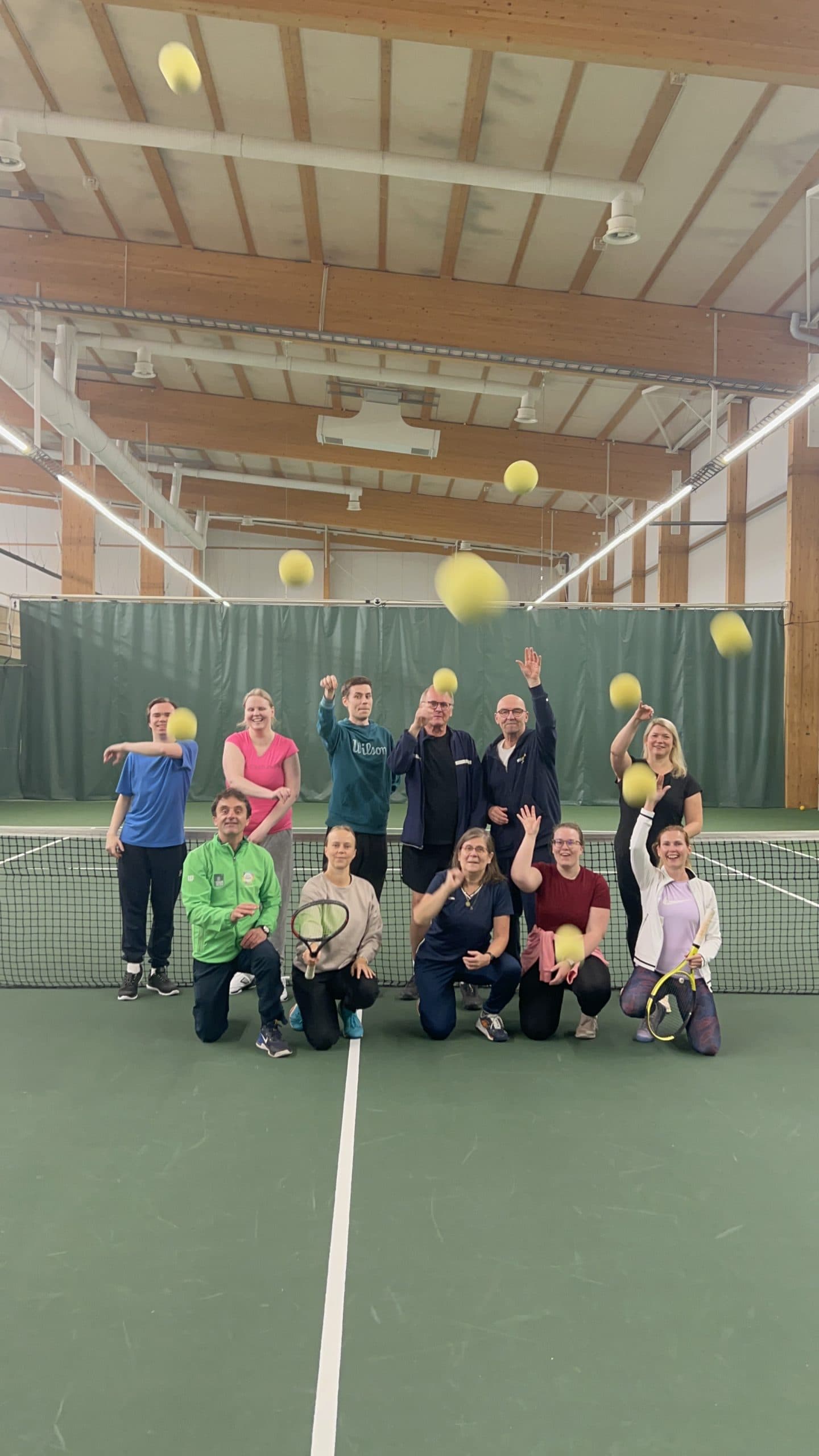 Henkilöitä tenniskentällä verkon edessä kahdessa rivissä. Osalla pelaajista tennismailat kädessä ja kaikki heittävät tennispallot kohti kameraa. Lähestyviä tennispalloja näkyy joka puolella kuvaa.